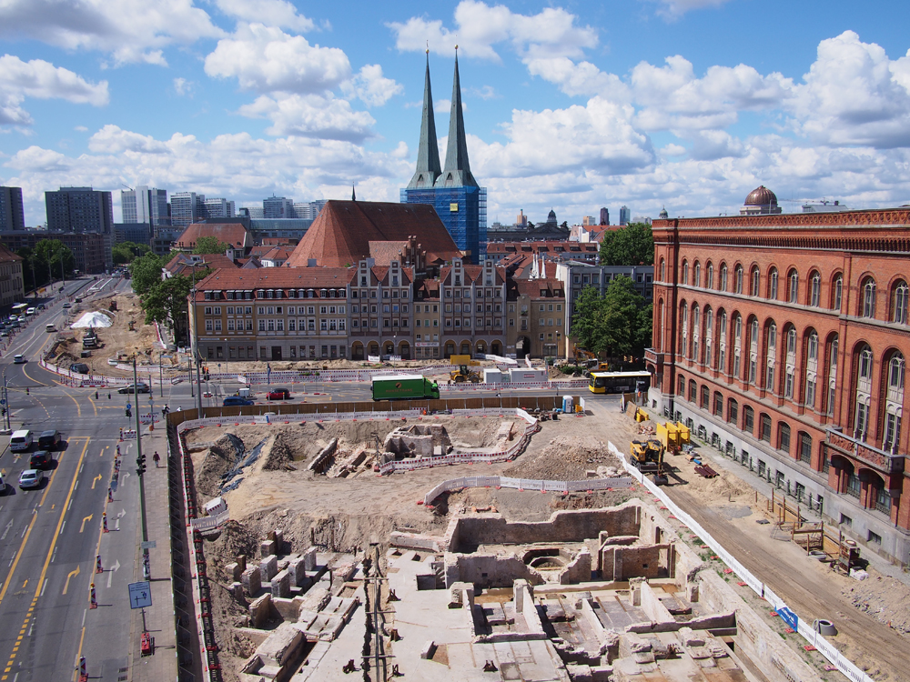 Blick auf die Ausgrabungsfläche am Berliner Molkenmarkt. Man sieht eine Vielzahl an freigelegten Keller- und Grundmauern. Rechts ist die Fassade des Roten Rathauses zu sehen und im Hintergrund die beiden Türme der Nikolaikirche, heute Museum.