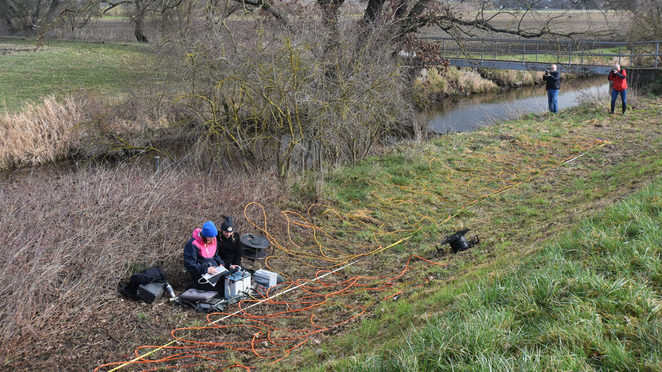 Zwei Frauen sitzen am Flussufer des Landgrabens vor einem Gerät, mit dem der elektrische Widerstand im Uferbereich gemessen wird. Dazu gehören zahlreiche verlegte Elektrokabel.